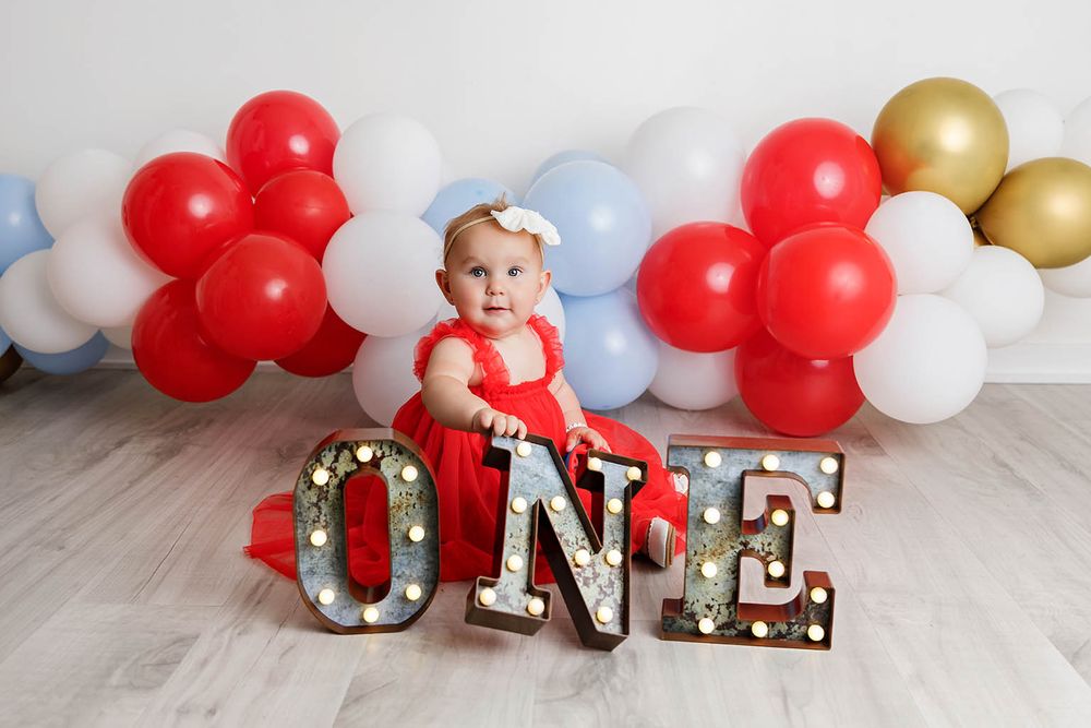 Baby girl in red dress sits on ONE marquee letters surrounded by red, white, blue, and gold balloons for first birthday.