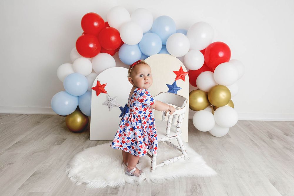 Toddler girl in floral dress poses by patriotic balloon arch with red, white, blue, and gold balloons and star decorations.