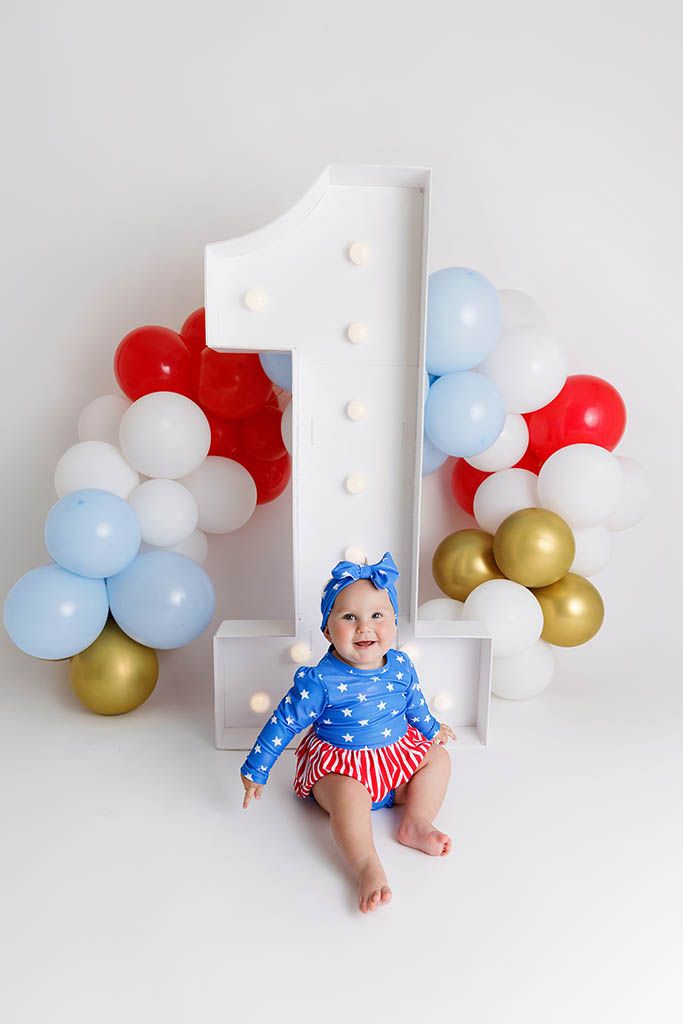 Baby girl in patriotic blue outfit sits before large number 1 with red, white, blue, and gold balloons for first birthday.