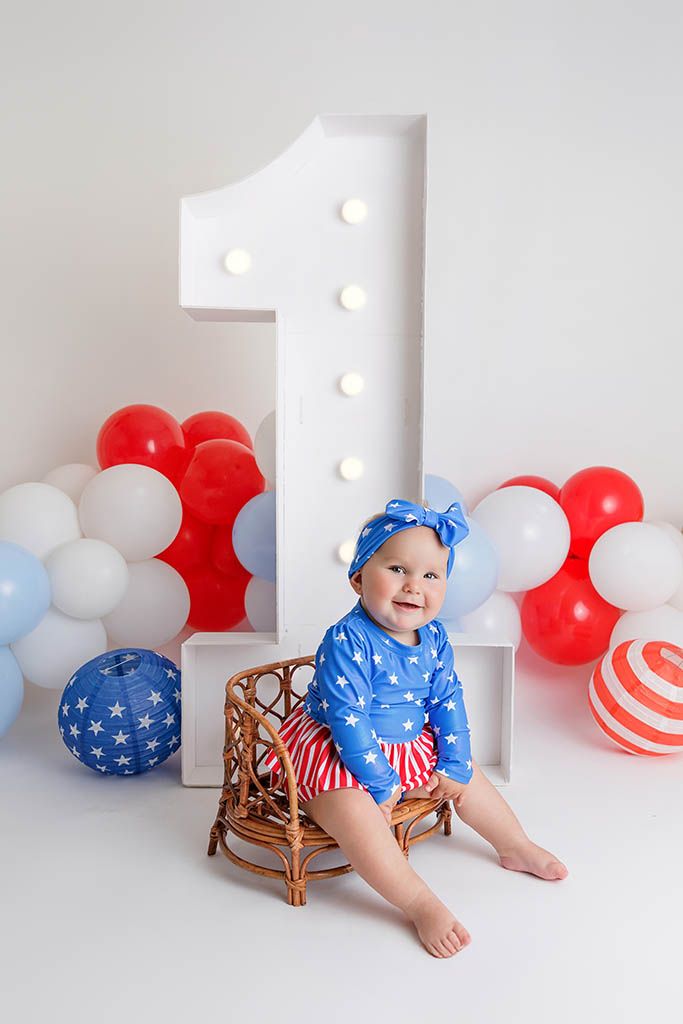Baby girl in patriotic star outfit by large number 1 with red white blue balloons, affordable baby photographer in Riverside county.