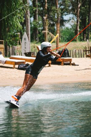 Wakeboarder riding cable park waves wearing helmet and wetsuit at sunny outdoor water sports venue.