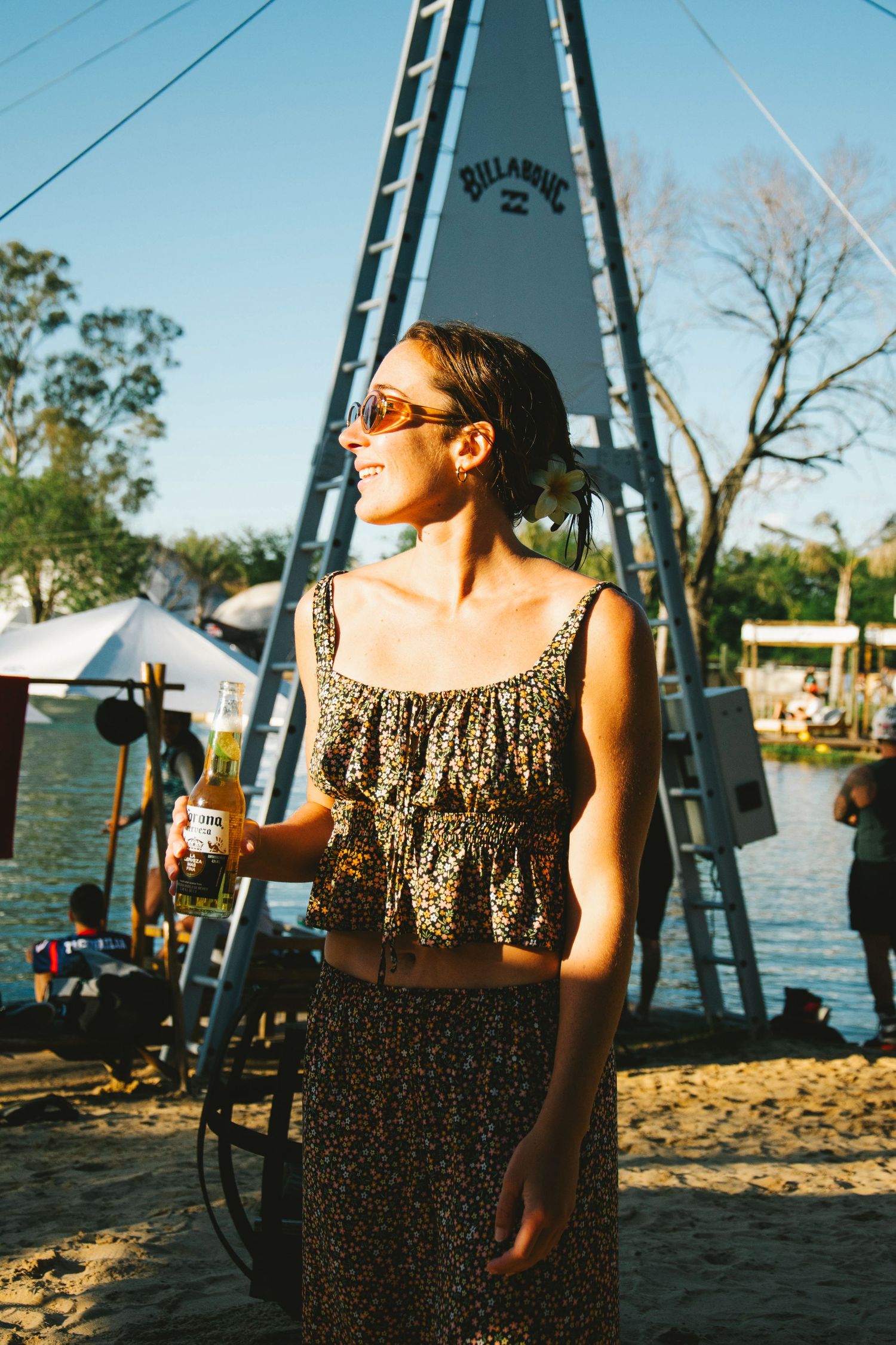 Woman in floral crop top and skirt holding a beer near a Billabong sign by a sunny lakeside.