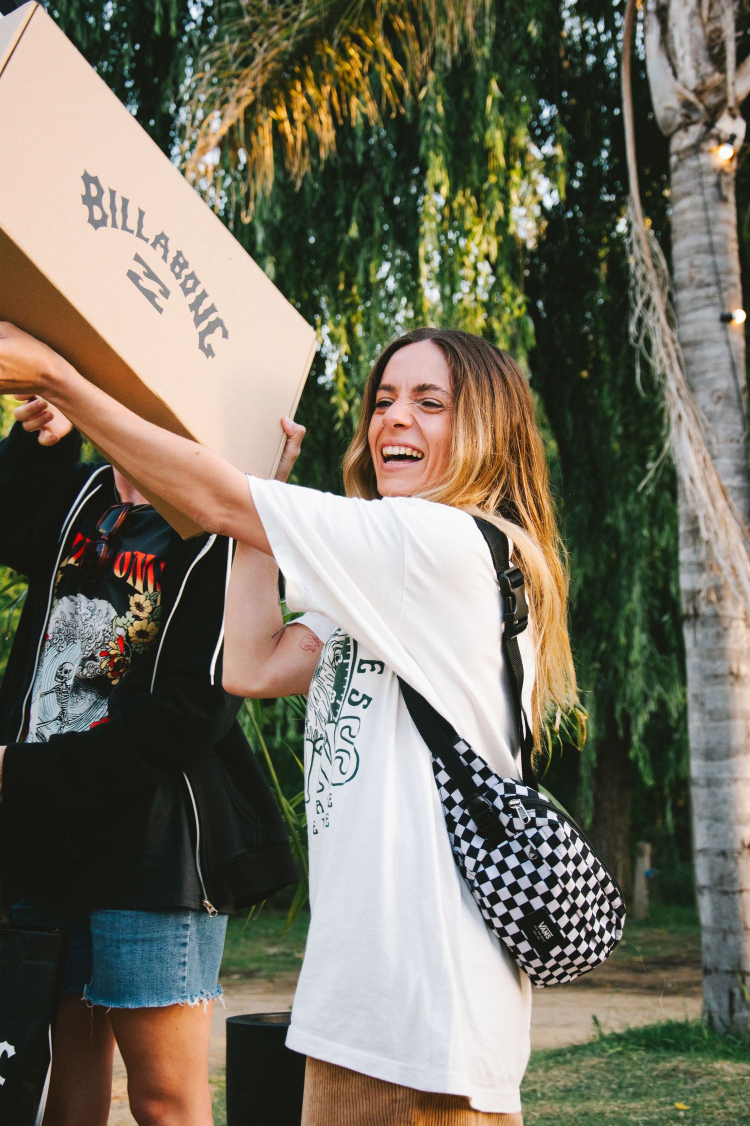 Woman laughing holding a Billabong box outdoors, wearing white graphic tee and checkered crossbody bag.