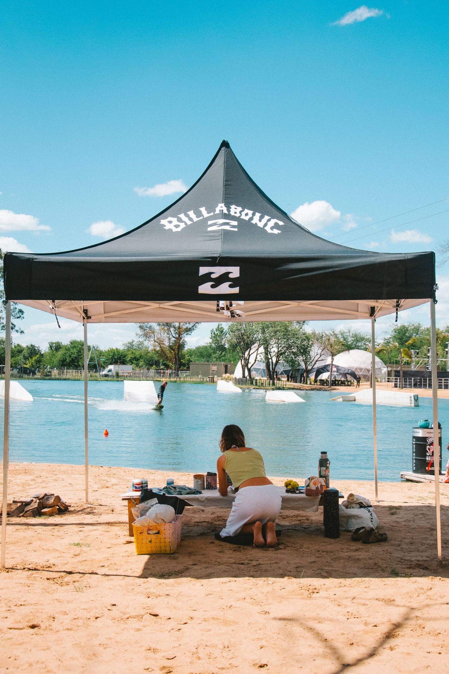 Person sitting under a black Billabong branded canopy tent beside a wakeboarding cable park lake.