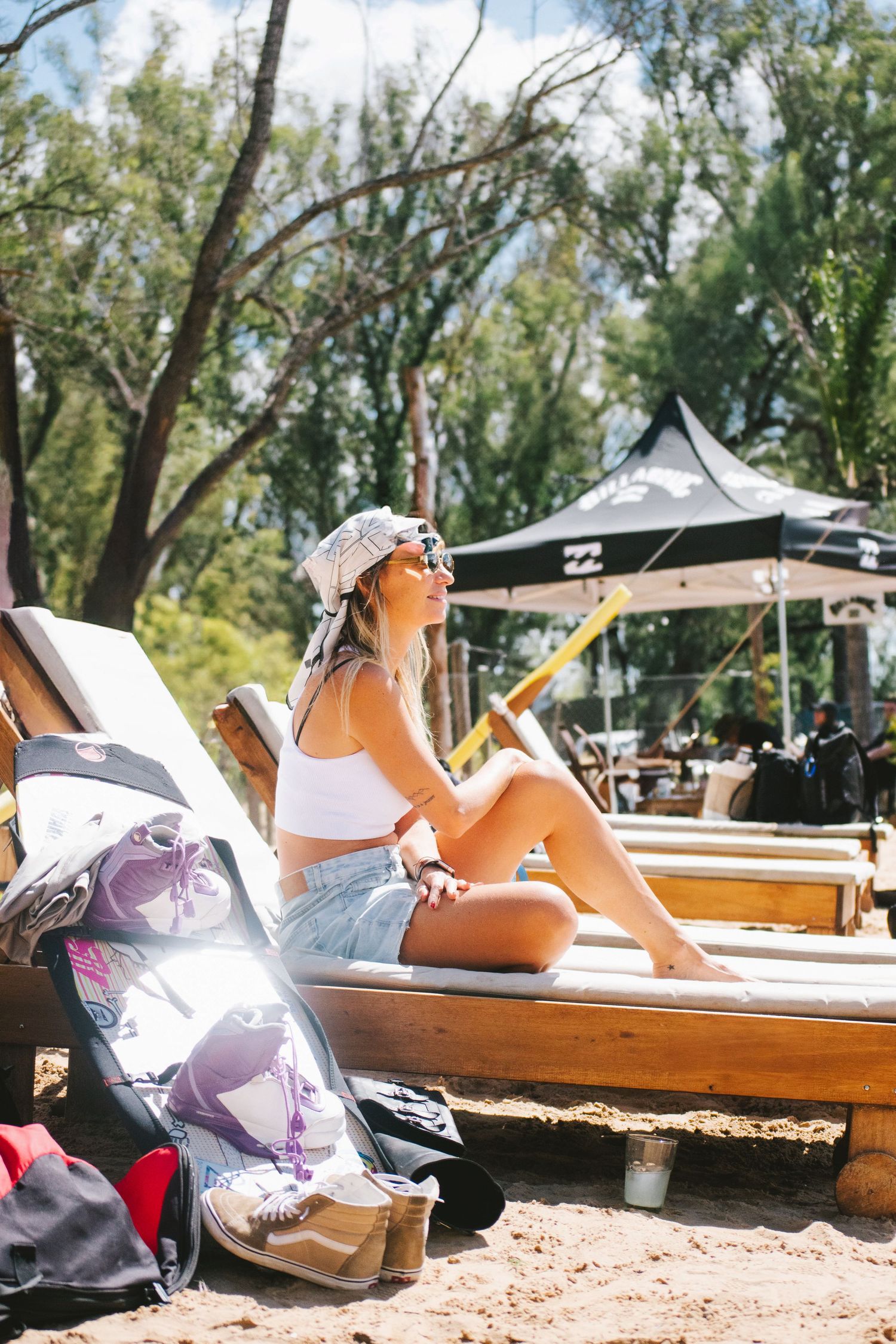 Young woman in white crop top and denim shorts relaxing on wooden beach lounger at outdoor event.