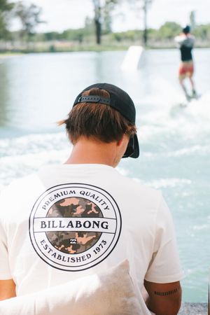 Man in Billabong t-shirt watching a wakeboarder on a lake.
