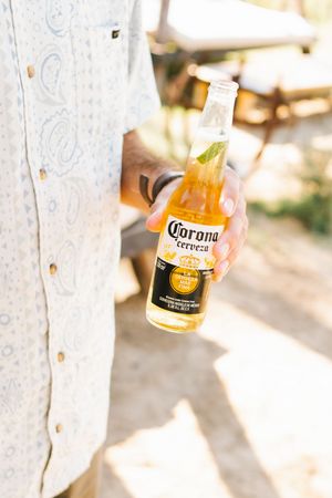Hand holding a Corona Cerveza beer bottle outdoors in bright sunlight.