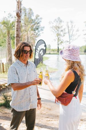 Man and woman toasting beer bottles near a Billabong flag at a lakeside outdoor festival.