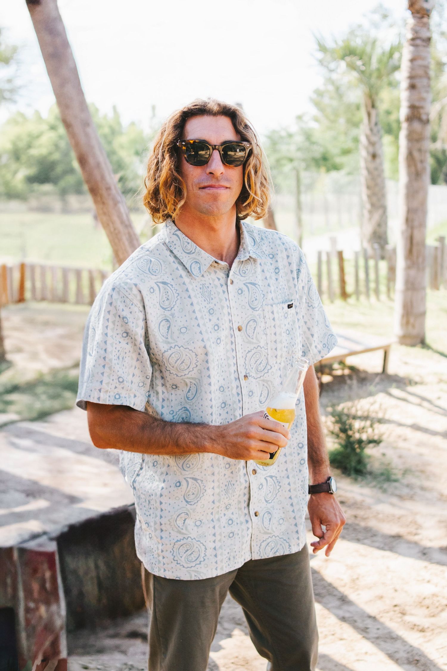 Surfer man with long hair wearing a paisley shirt and sunglasses holding a beer outdoors.