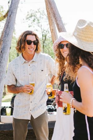 Group of friends in sunglasses smiling and holding beers at a sunny outdoor event.