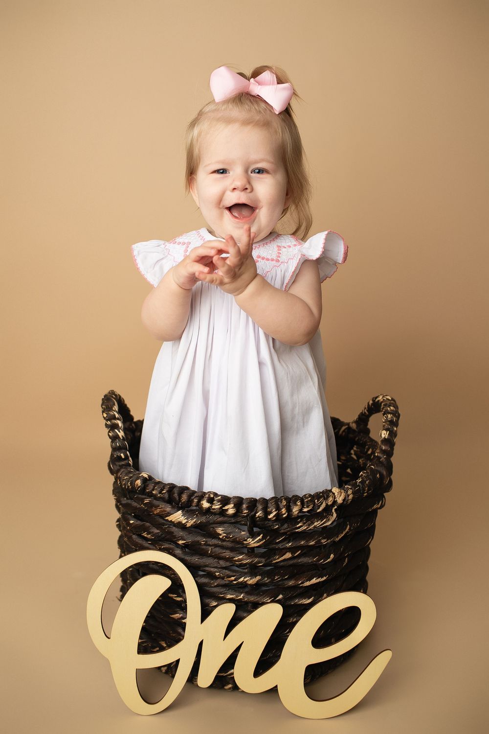 Laughing baby girl in white dress and pink bow stands in wicker basket beside wooden One sign.