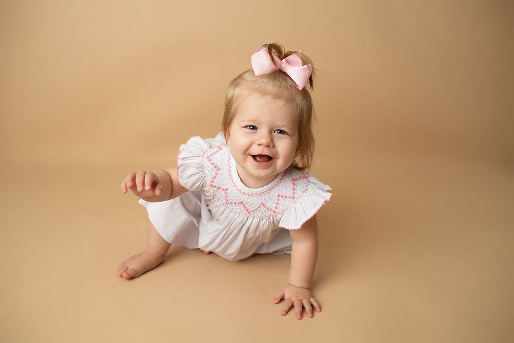Happy baby girl in white smocked dress and pink bow crawls on beige background during milestone session.