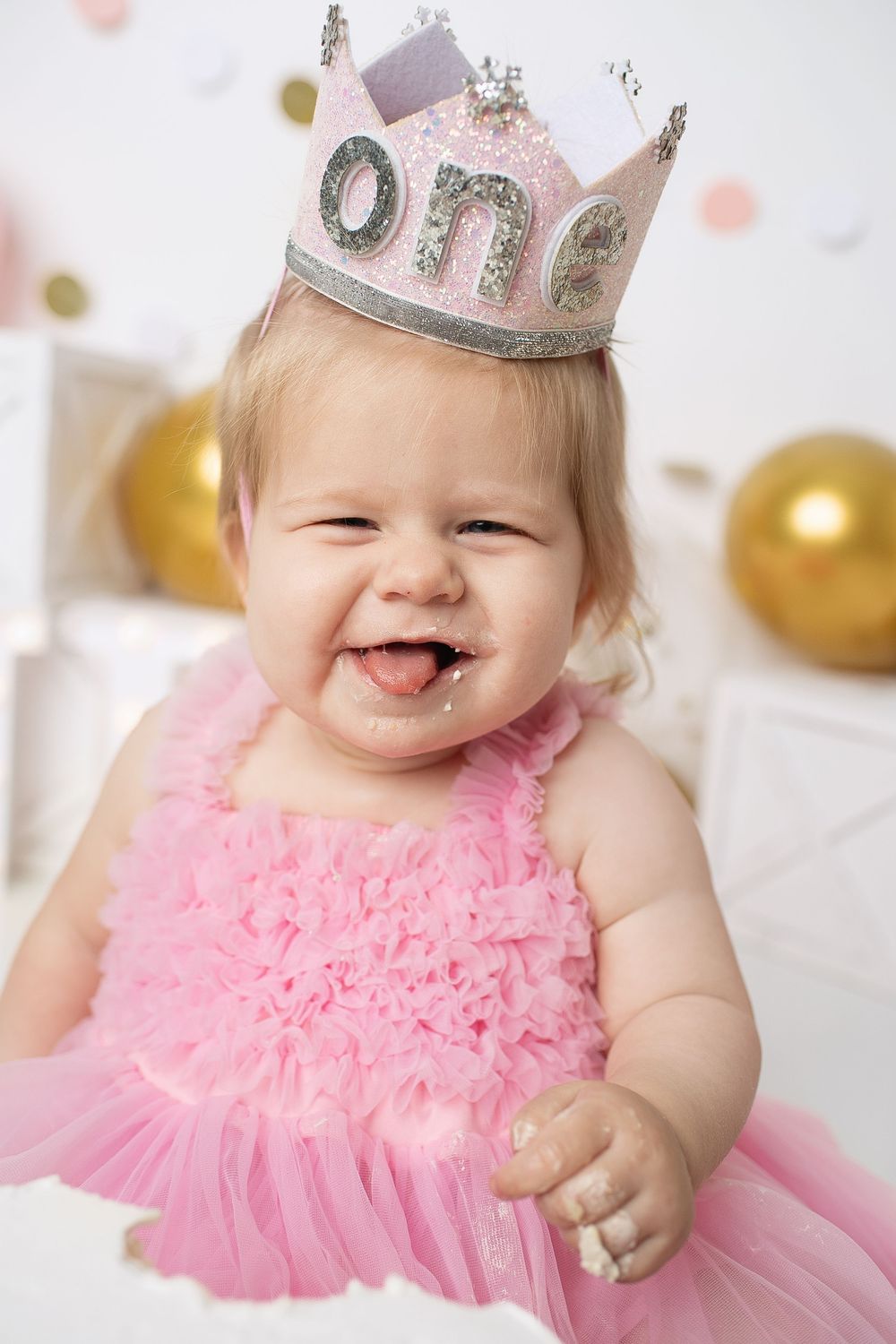Joyful baby girl in pink tutu dress wearing glittery One crown laughs during first birthday cake smash.