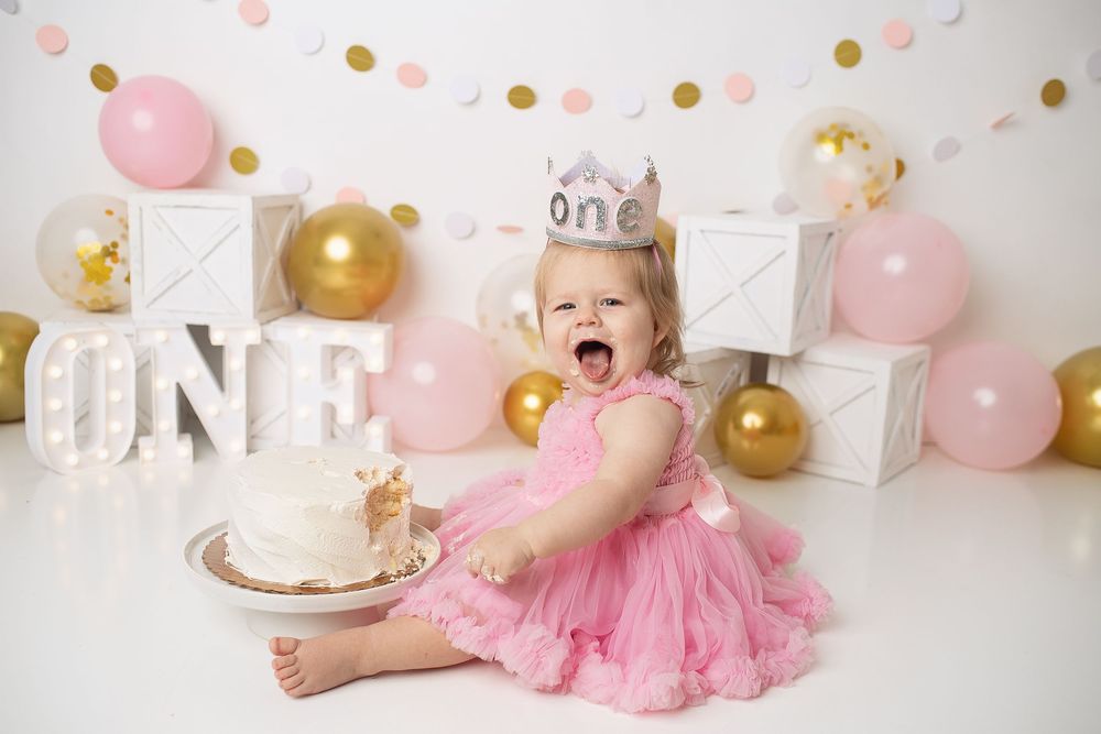 Excited baby girl in pink dress and One crown reaches for cake during first birthday cake smash session.