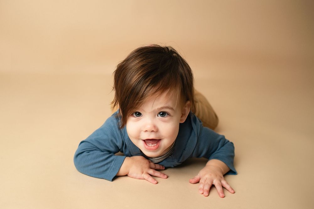 Smiling baby boy in blue outfit lies on beige background during milestone photography session.