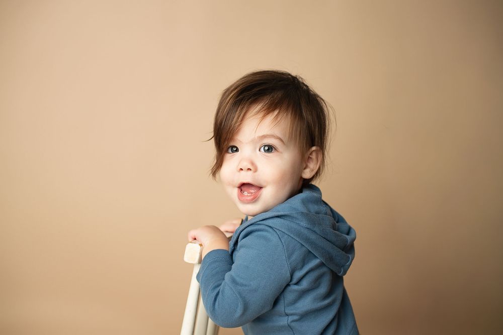 Happy toddler in a blue hoodie leaning on a white chair, smiling over shoulder on beige background.