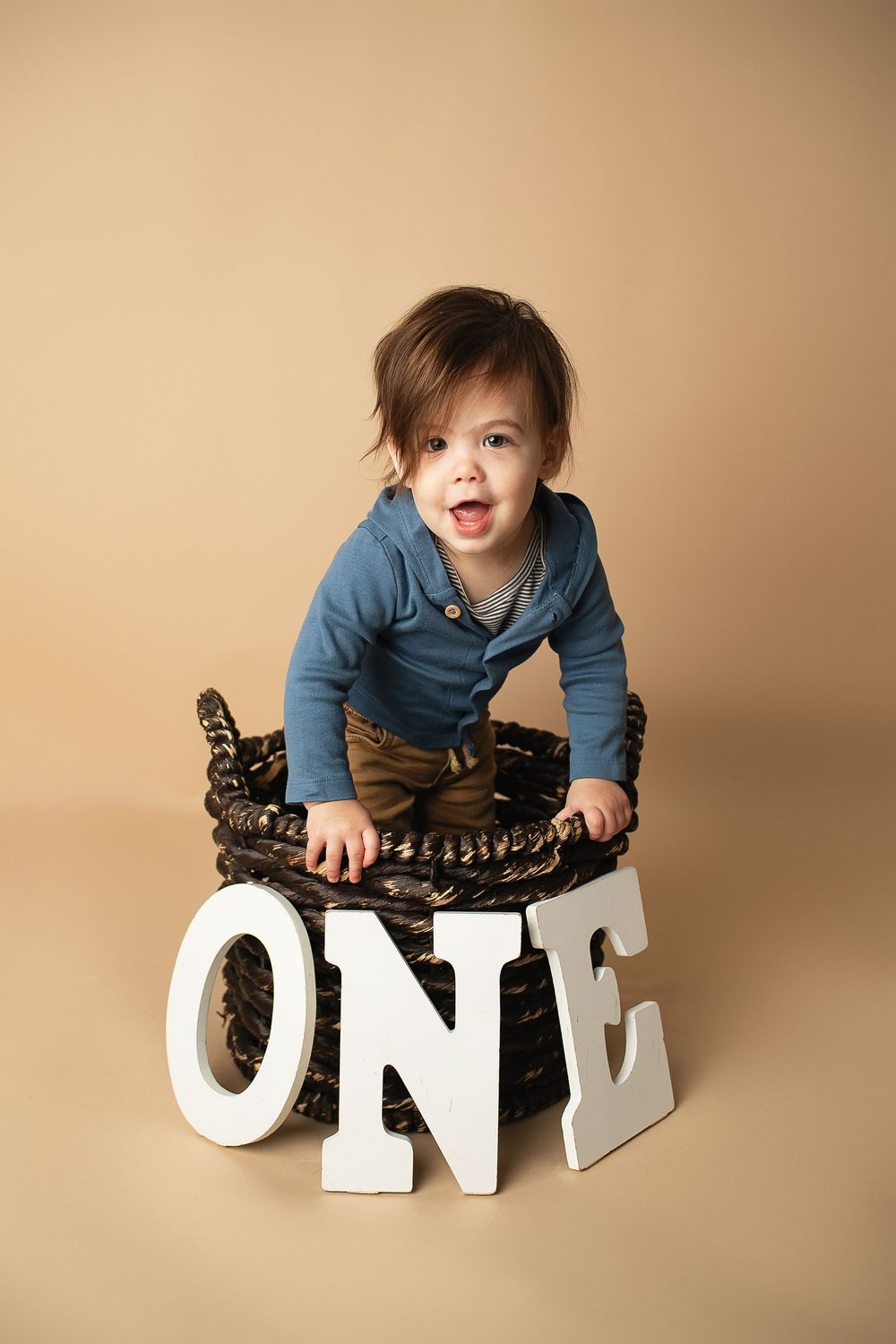 Cheerful one-year-old baby in blue shirt sitting in a wicker basket with ONE letter props.