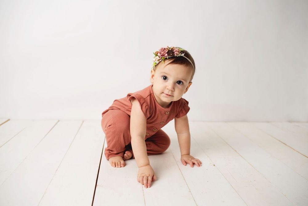 Baby girl in rust romper and floral headband crawling on white wood floor.
