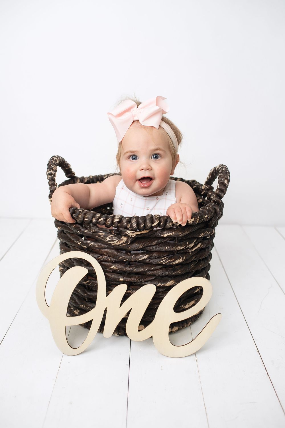 Baby girl in white dress and pink bow sits in wicker basket beside wooden One sign for first birthday photo.