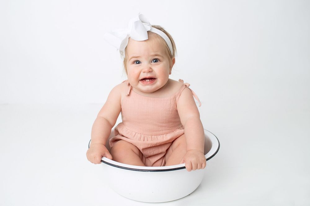 Smiling baby girl in pink romper and white bow headband sits in white bowl for milestone photo.