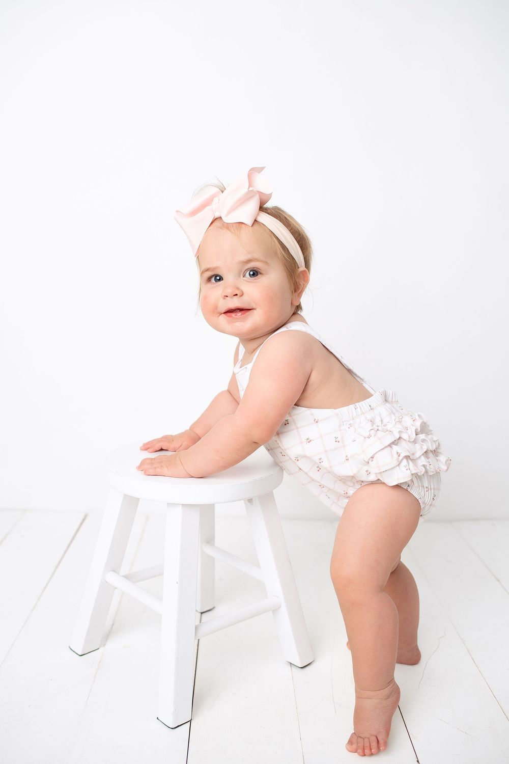 Smiling baby girl in white romper and pink bow standing at white stool.
