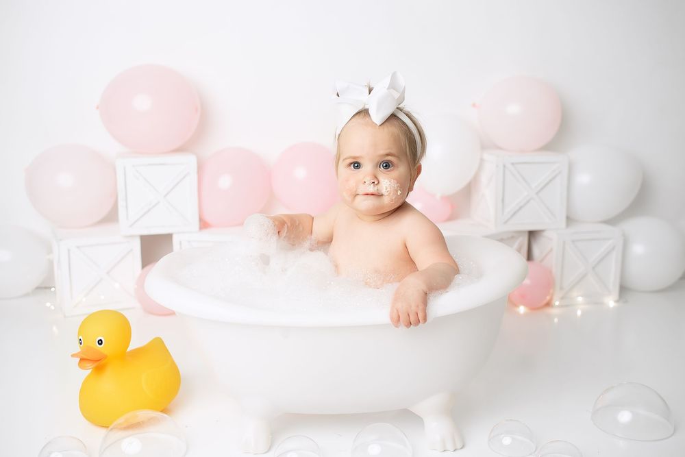Baby girl in white bow sits in bubble bath tub with rubber duck during first birthday photo session.