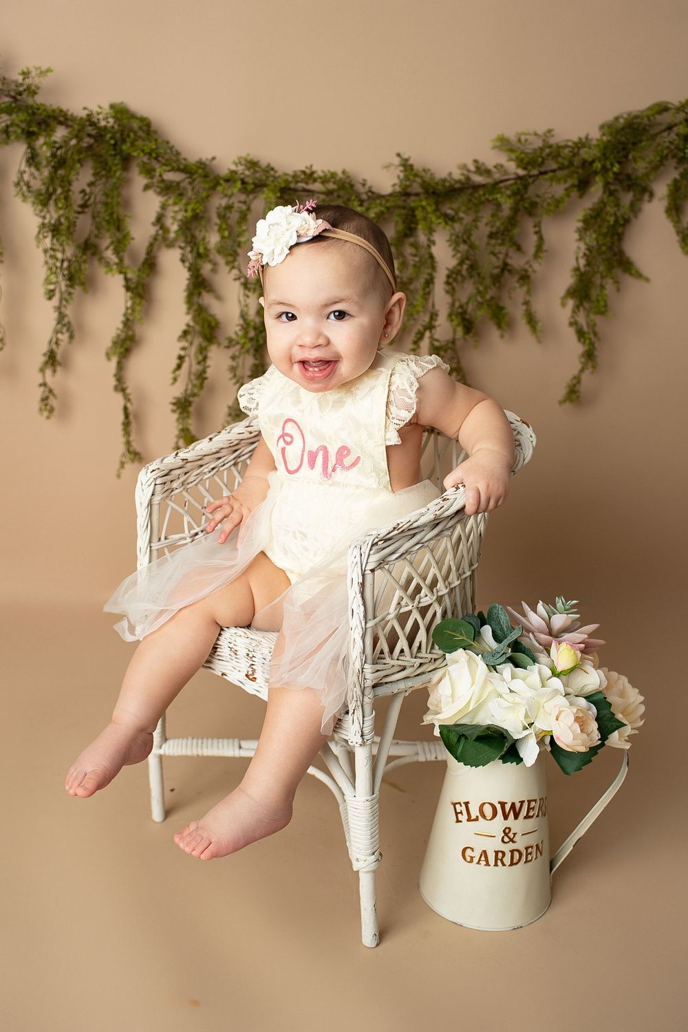 Laughing baby girl in a white 'One' romper sitting in a wicker chair with garden flowers for her birthday.