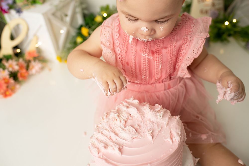 Baby girl in a pink dress smashing her first birthday cake covered in pink frosting during photo session.