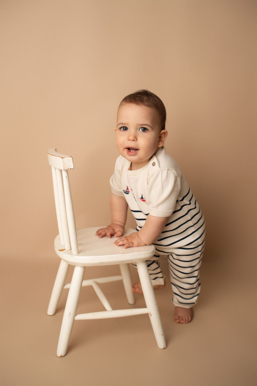Baby in white striped romper standing beside a small white chair on a beige background.