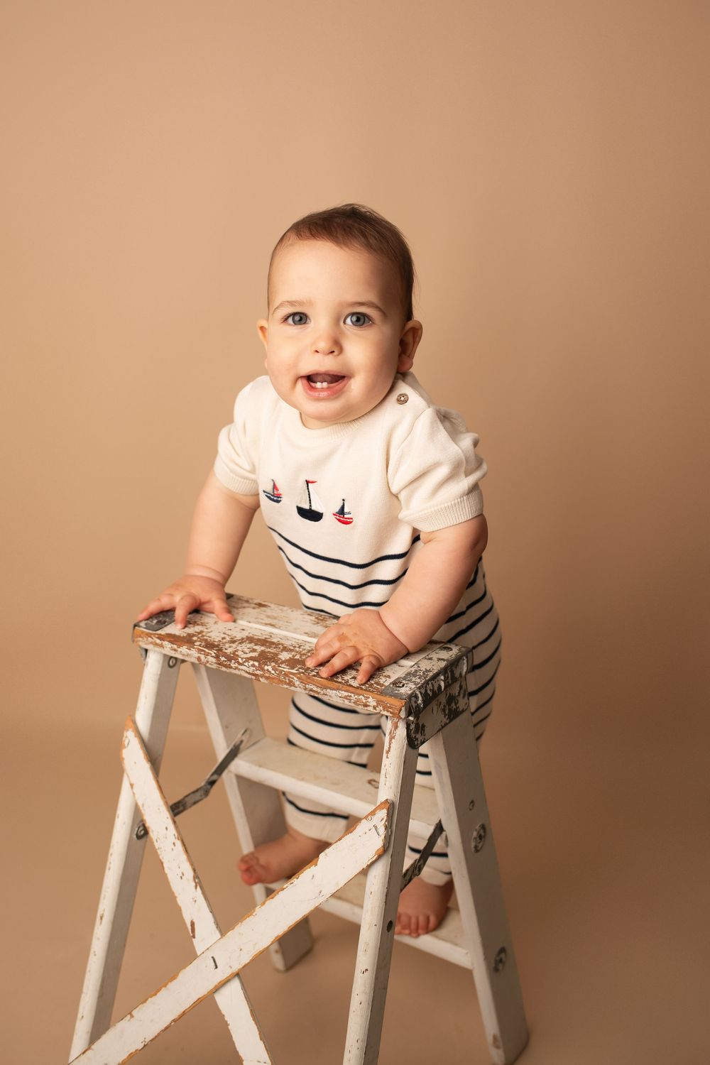 Smiling baby in nautical striped romper standing on a rustic white step ladder on beige background.