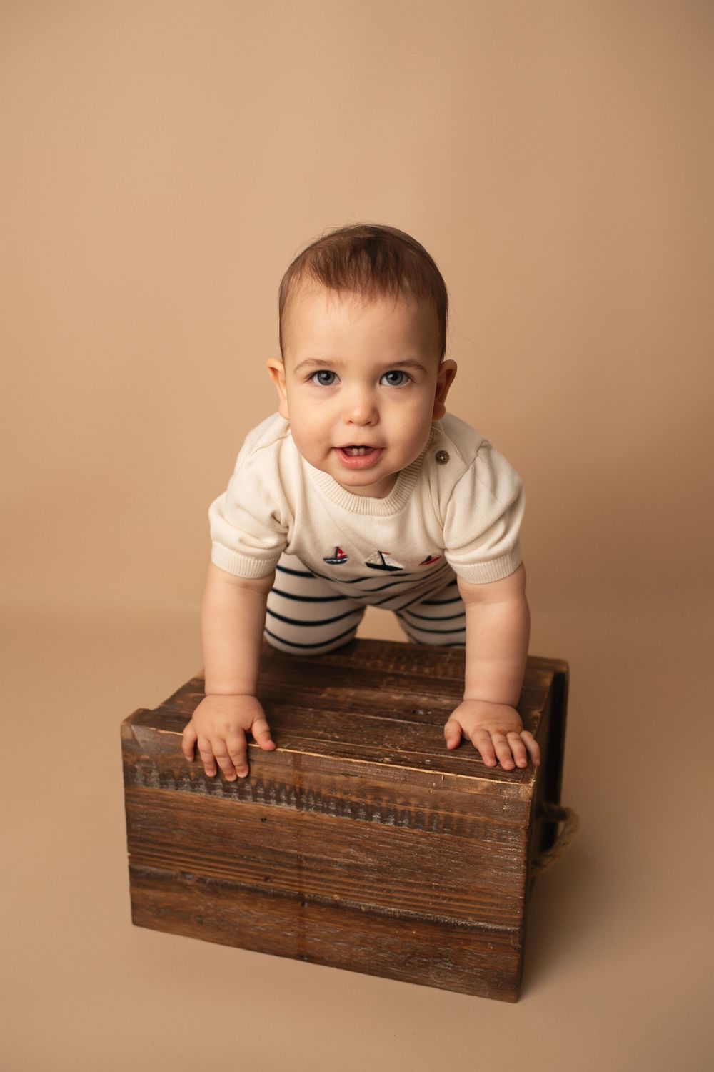 Cute baby in white striped onesie leaning over a rustic wooden crate on a beige background.