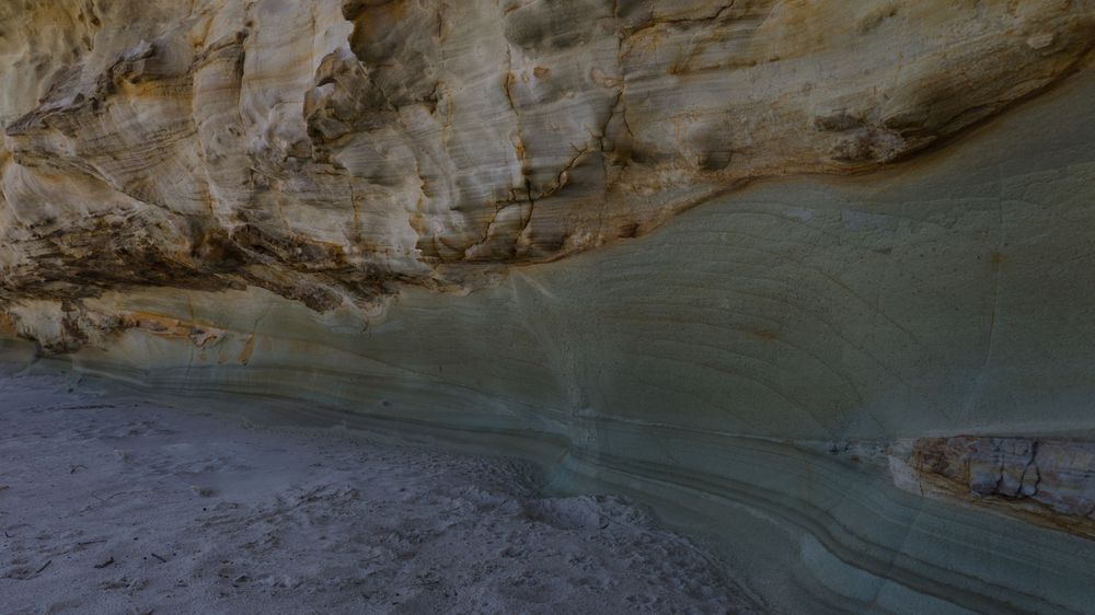Colorful layered rock cave wall with turquoise and cream mineral deposits along a sandy beach floor.