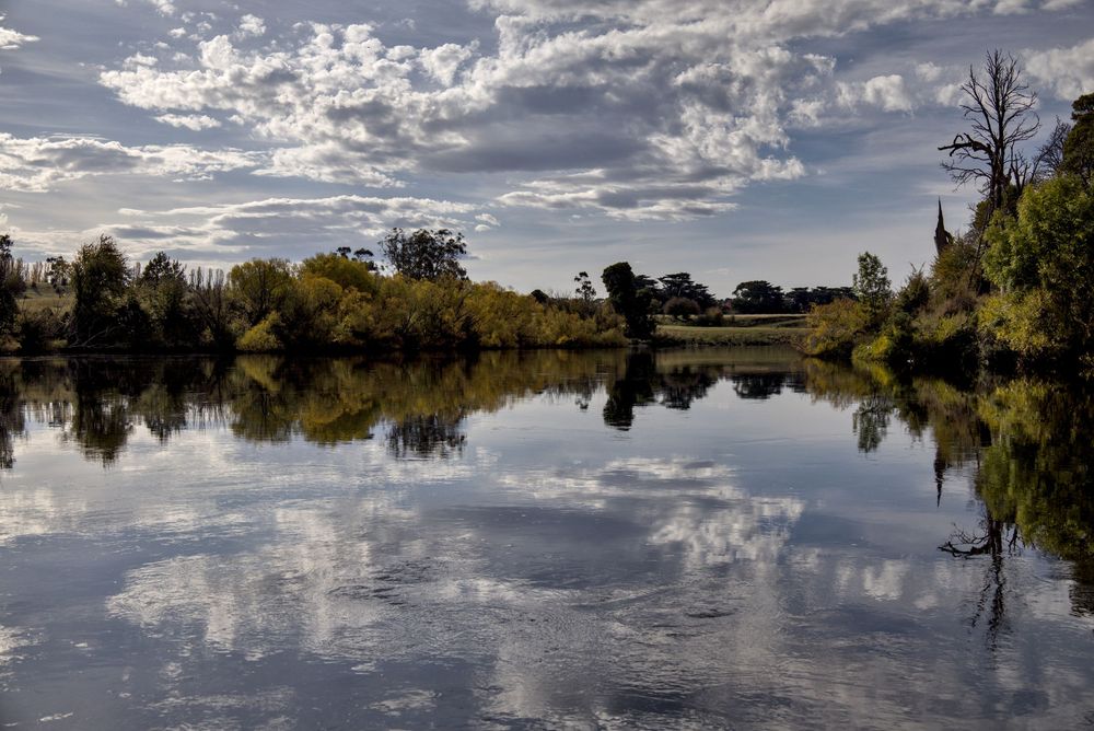 Calm river reflecting clouds and autumn trees on a peaceful overcast day.