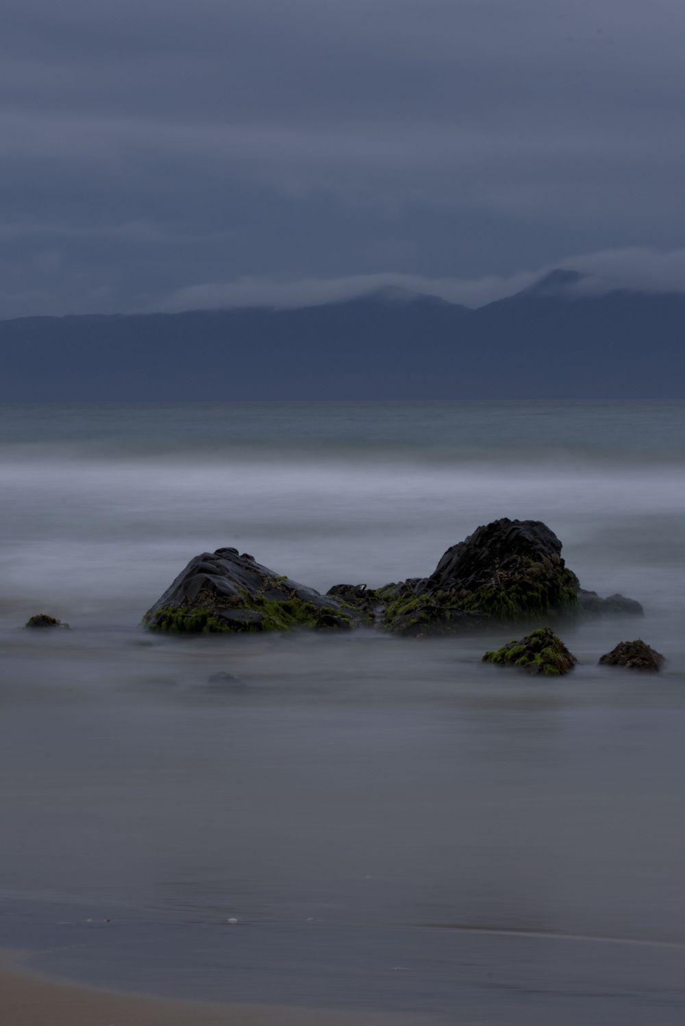 Moody seascape with mossy rocks on a misty beach, calm ocean waves, and distant mountains under overcast skies.