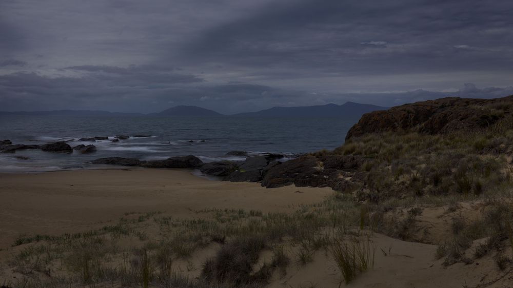 A moody coastal beach scene at dusk with dark clouds, rocky shores, grassy dunes, and distant mountains across the sea.