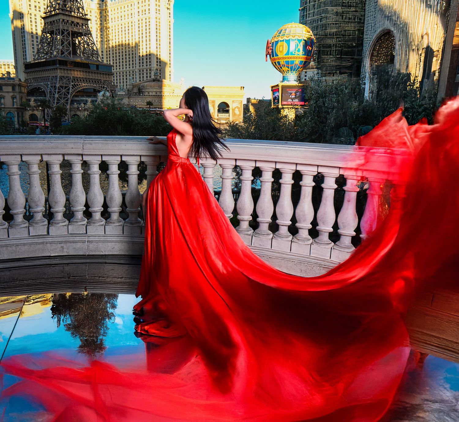 Woman in flowing red gown poses on balcony with hot air balloon and Las Vegas skyline backdrop.