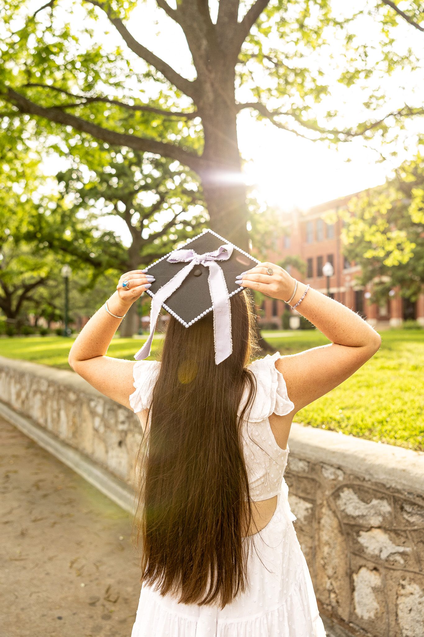 Graduate holds mortarboard cap outdoors in golden sunlight on a lush college campus.
