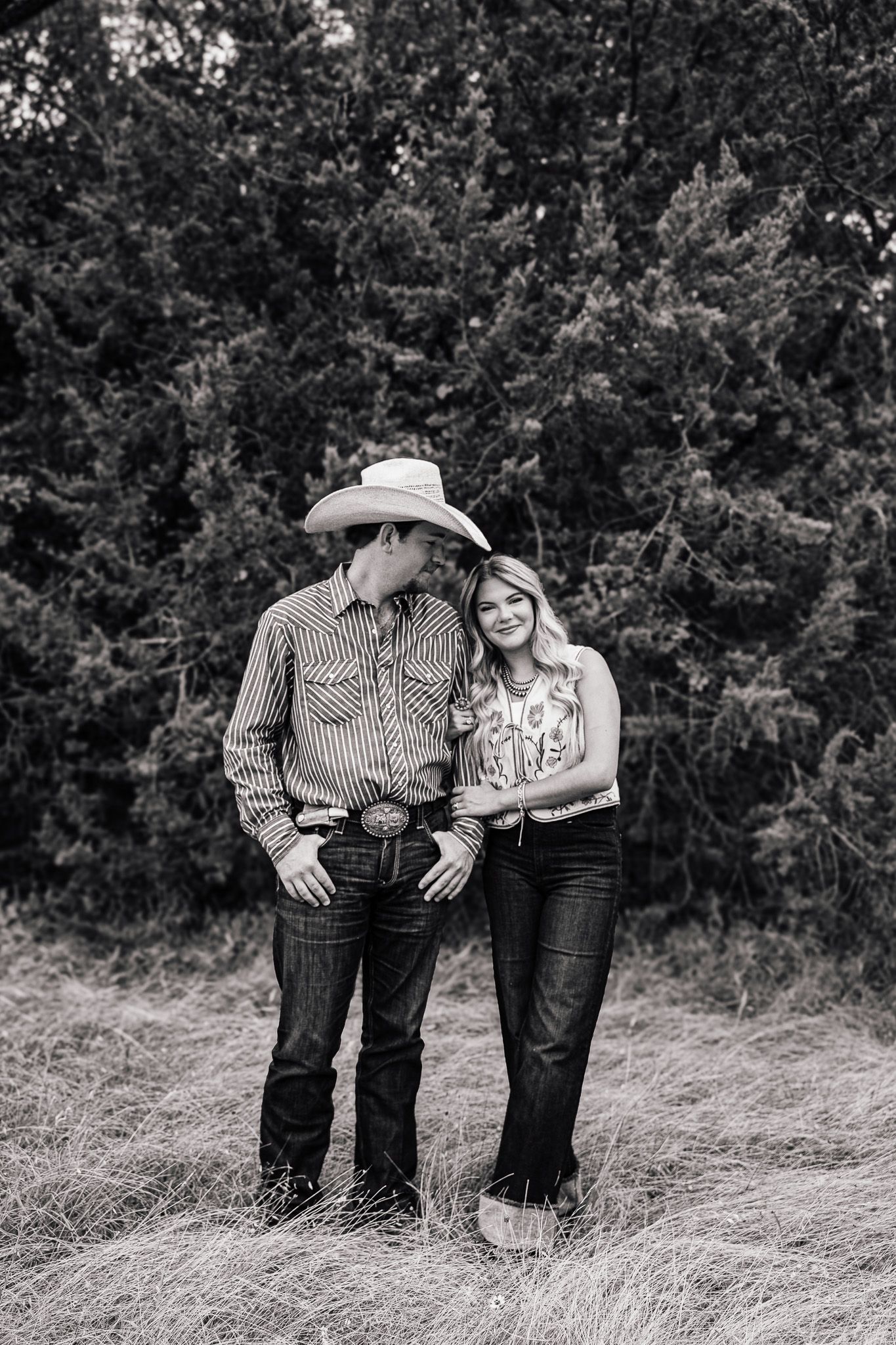 Black and white photo of a couple in western wear, man in cowboy hat and striped shirt, woman smiling, standing in a field.