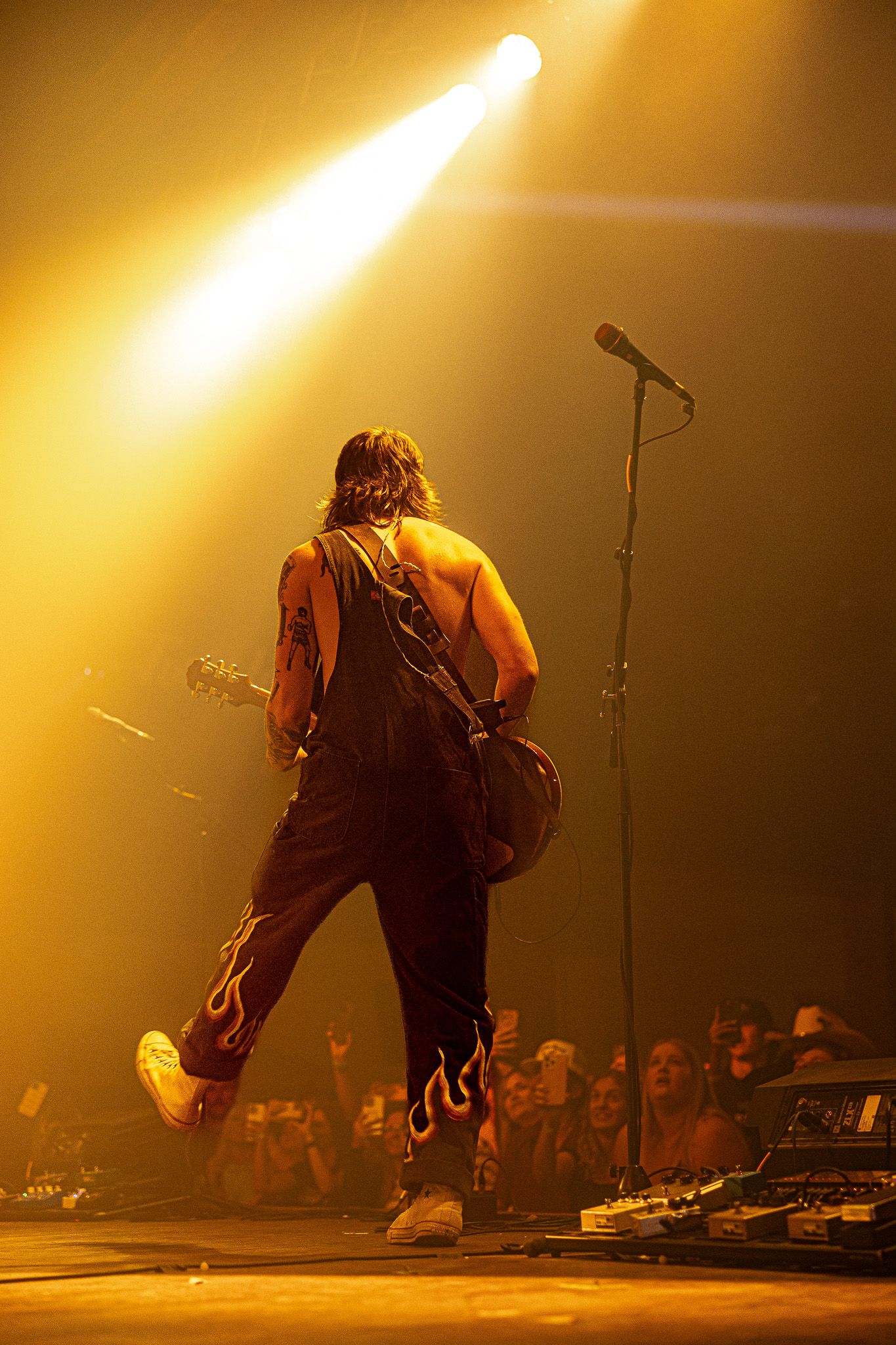 Shirtless rock musician in flame overalls jumps on stage with guitar under dramatic yellow spotlight.