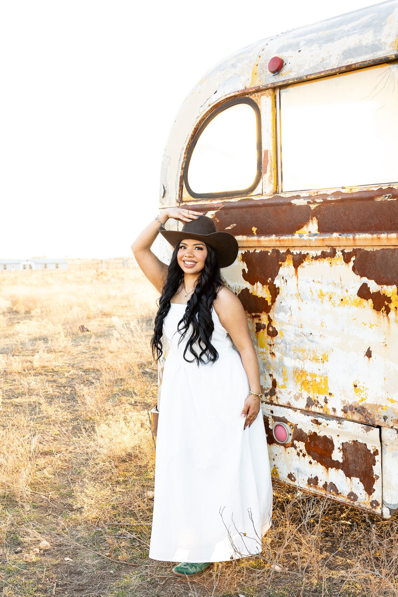 Smiling woman in white dress and cowboy hat poses beside a rusty vintage bus in a field.
