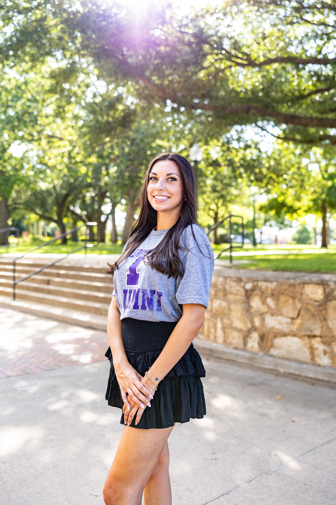 Smiling young woman in a purple alumni t-shirt poses on a sunny college campus walkway.