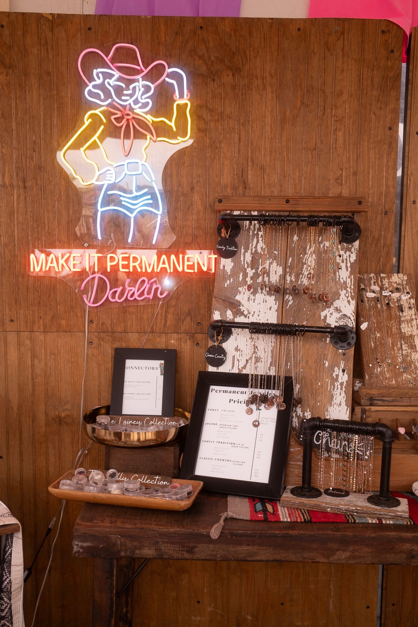 Neon cowgirl sign reading Make It Permanent Darlin above a rustic jewelry display table with framed menus and accessories.
