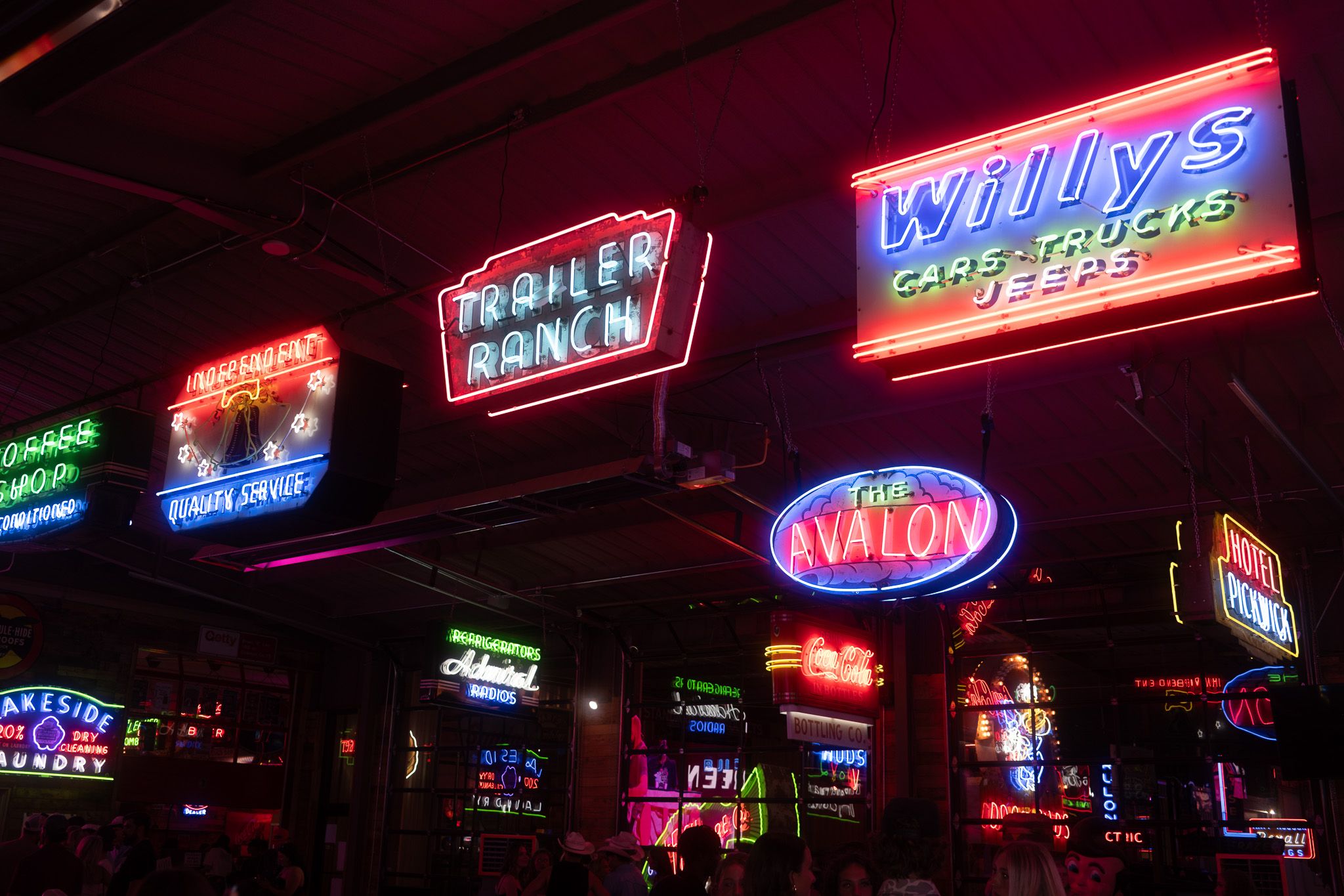 Colorful vintage neon signs including Willy's Cars and The Avalon displayed on a dark ceiling in a retro bar.
