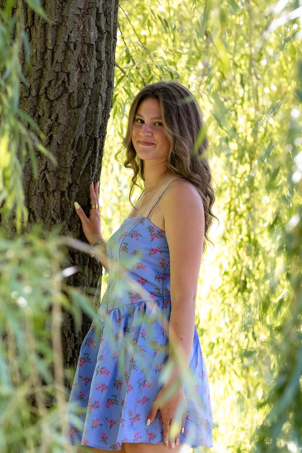 Young woman in blue floral sundress smiling beside a tree, surrounded by green willow branches outdoors.