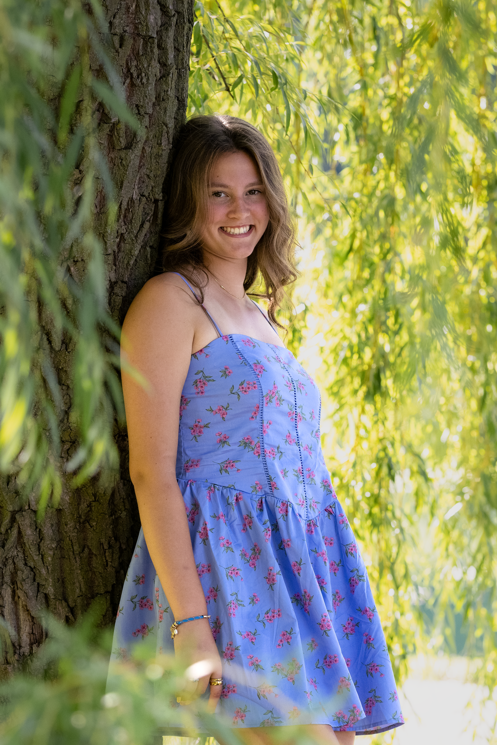 Young woman in blue floral sundress smiling outdoors leaning against a tree surrounded by green willow leaves.