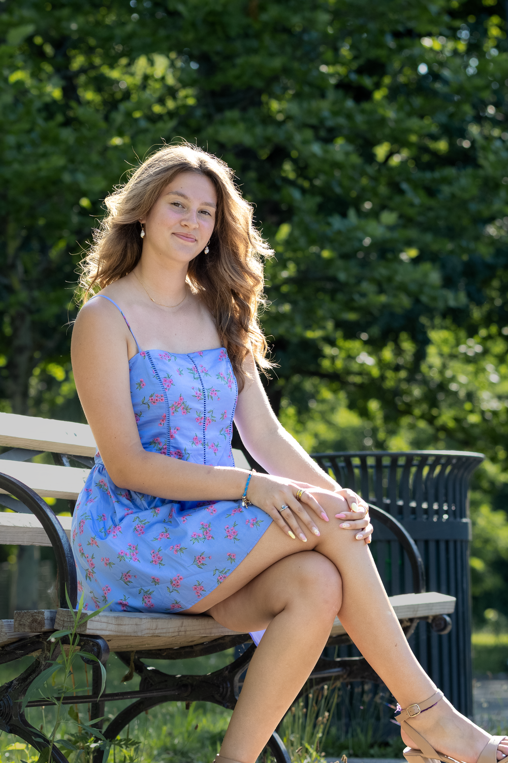 Young woman in blue floral sundress sitting on a park bench, smiling outdoors with green trees in background.