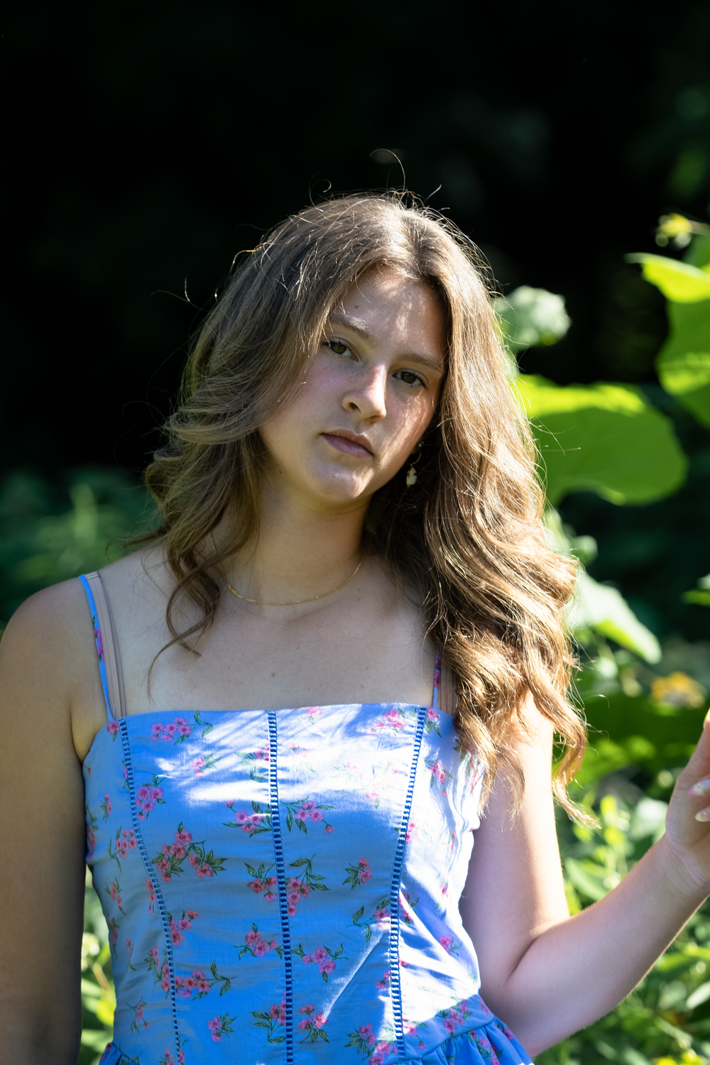 Young woman in blue floral sundress posing outdoors among lush green foliage in natural sunlight.