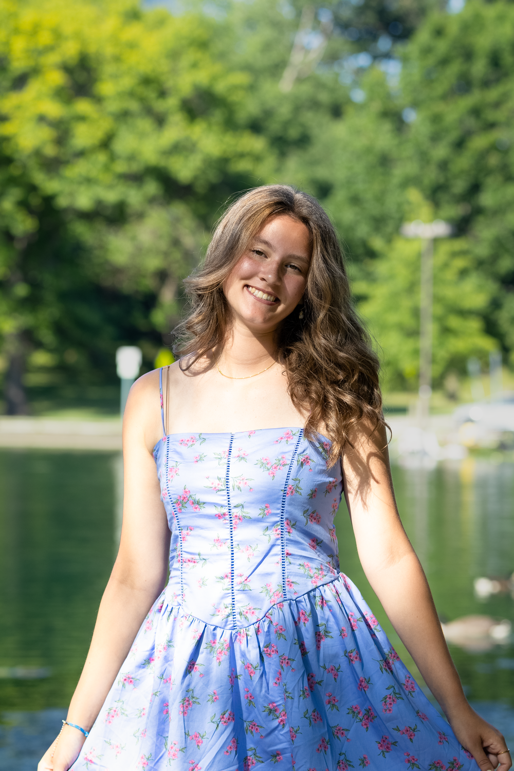 Smiling young woman in a blue floral sundress standing outdoors near a lake surrounded by green trees.