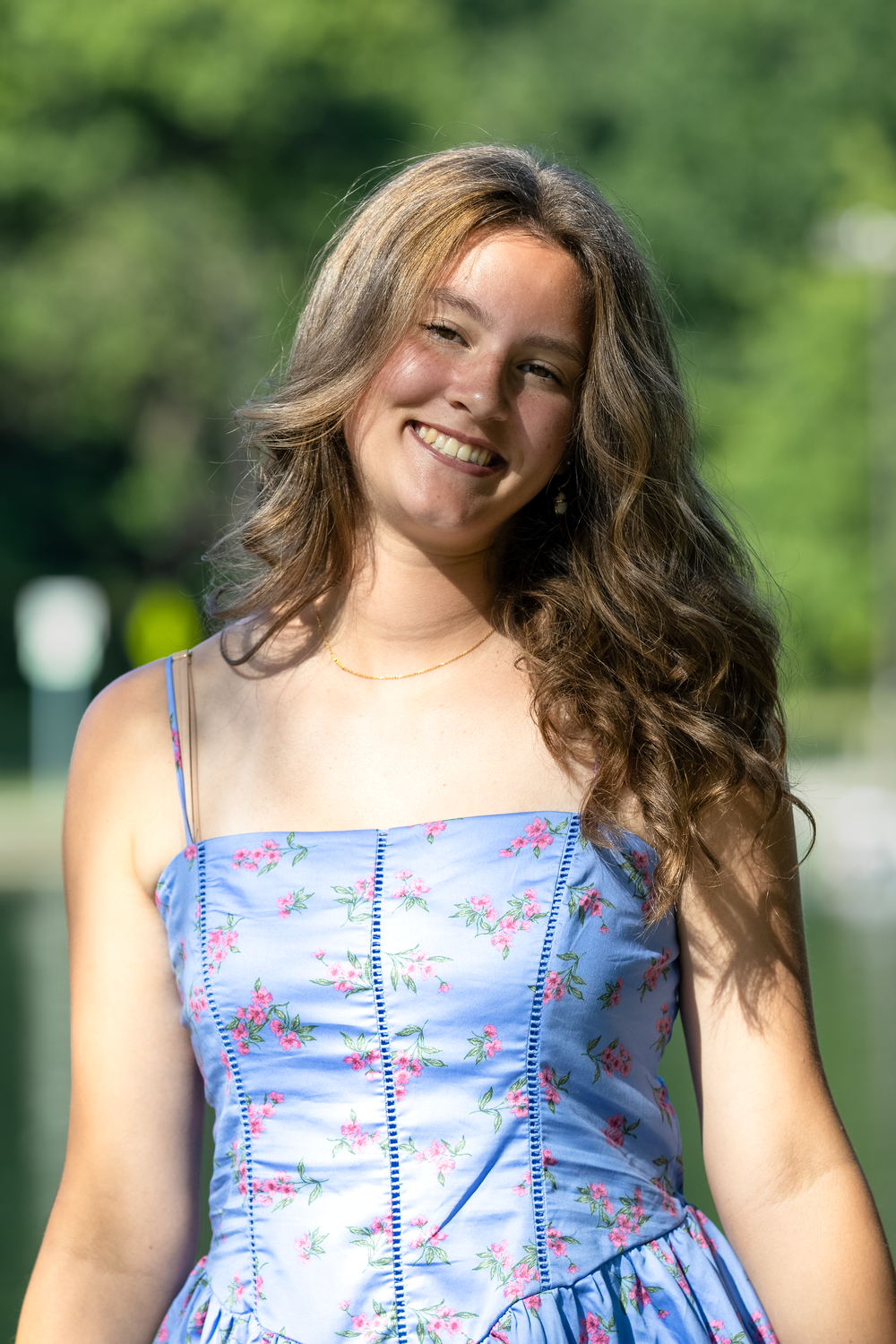 Smiling young woman with long wavy hair wearing a blue floral sundress outdoors in a sunny park setting.