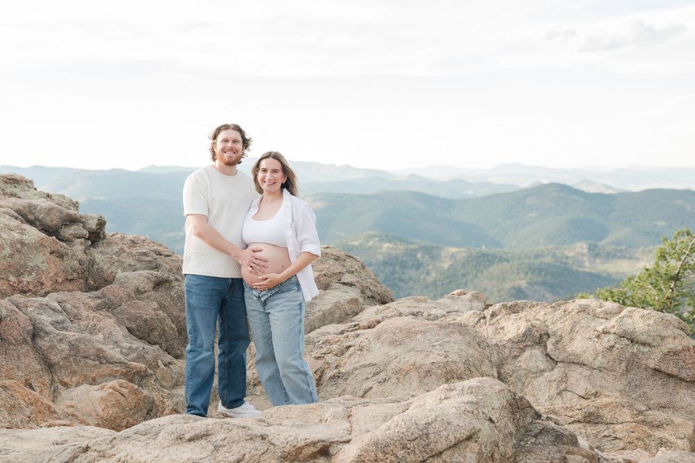 Couple poses on rocky mountain summit with sweeping valley views during maternity photo session outdoors.